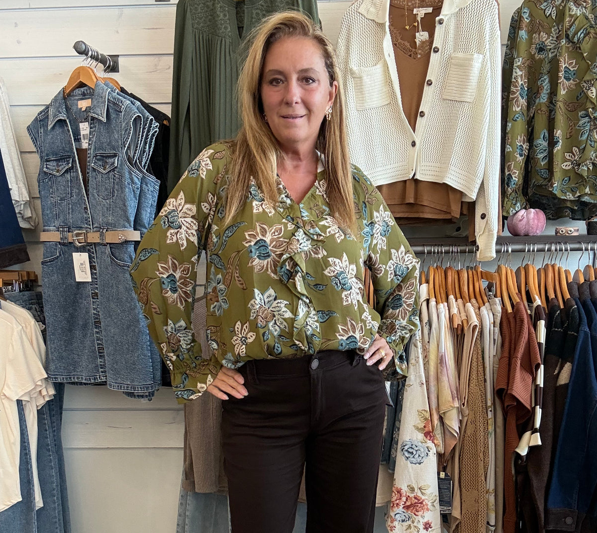 A woman stands in a clothing boutique wearing a sage green floral long-sleeve blouse and dark brown cropped pants. Clothing racks and shelves are visible behind her.