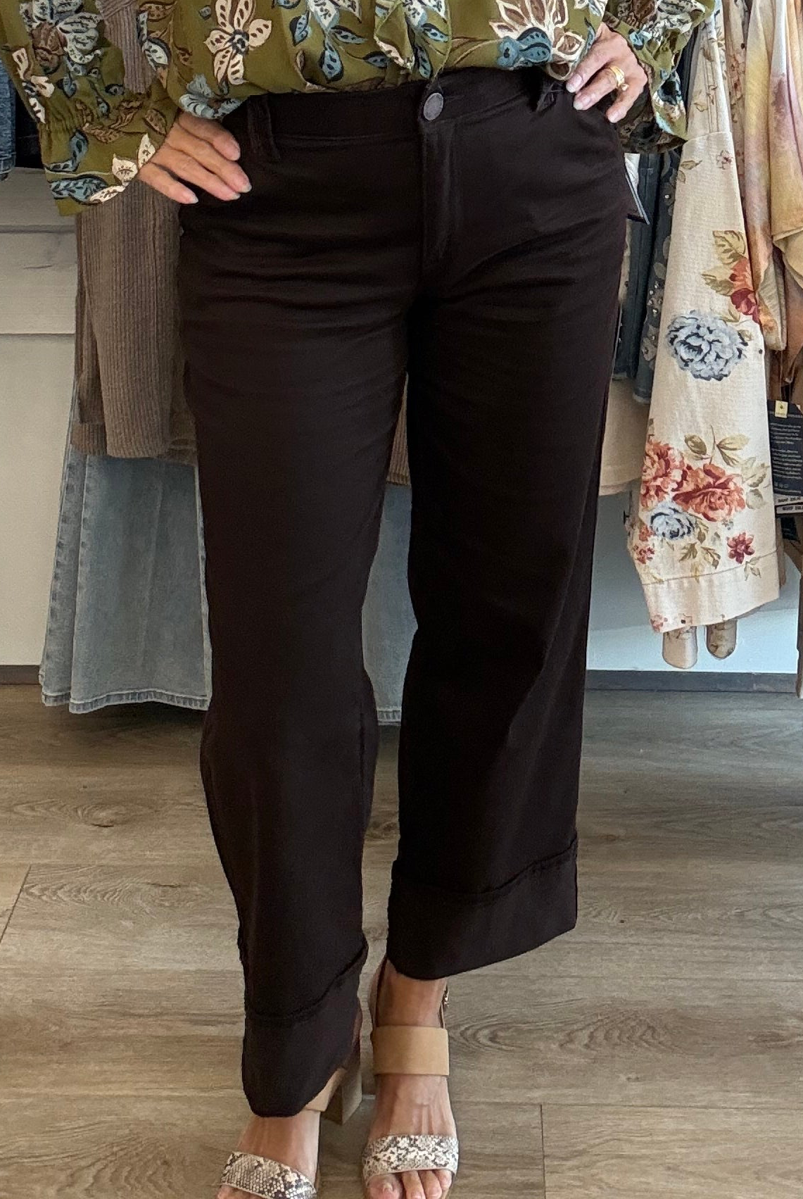 A person wearing dark brown cropped pants with rolled cuffs, paired with a floral blouse and beige heeled sandals, standing in a clothing store on a wood floor.