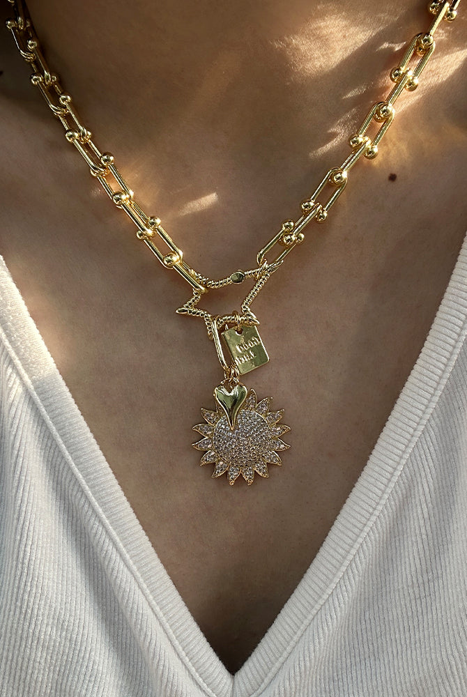 A close-up of a person wearing a gold chain necklace with a star-shaped connector and a sunburst pendant adorned with small clear stones, styled with a white ribbed V-neck top.