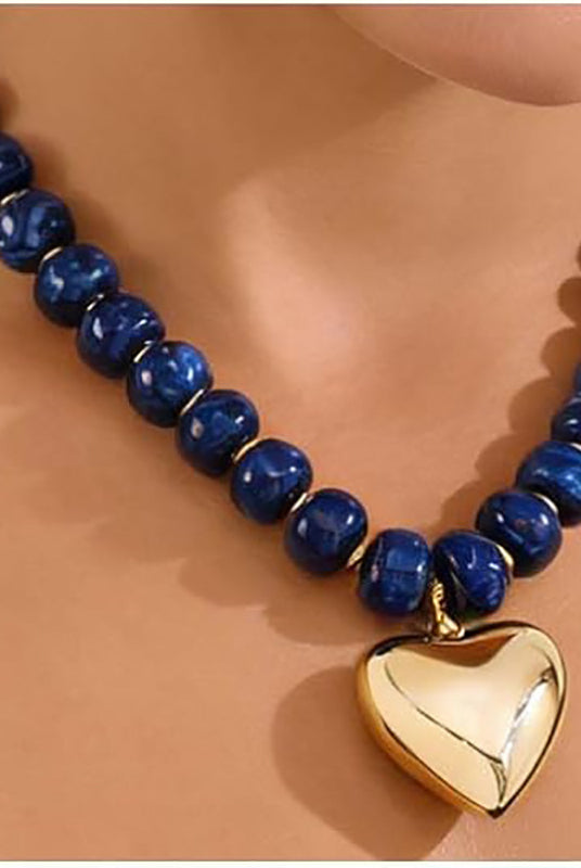 A beaded necklace with polished dark blue round beads and a shiny gold heart-shaped pendant, displayed on a mannequin neck against a warm beige background.