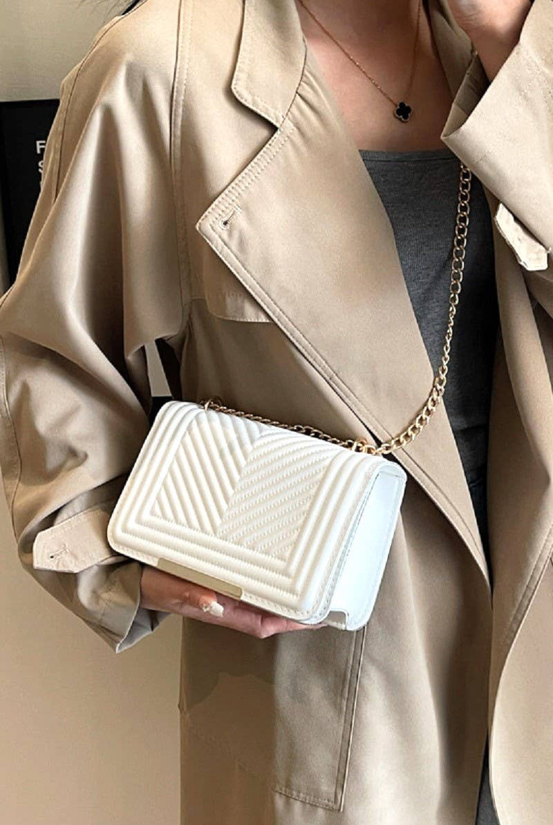 Person holding a white handbag with a gold chain strap, wearing a beige trench coat.