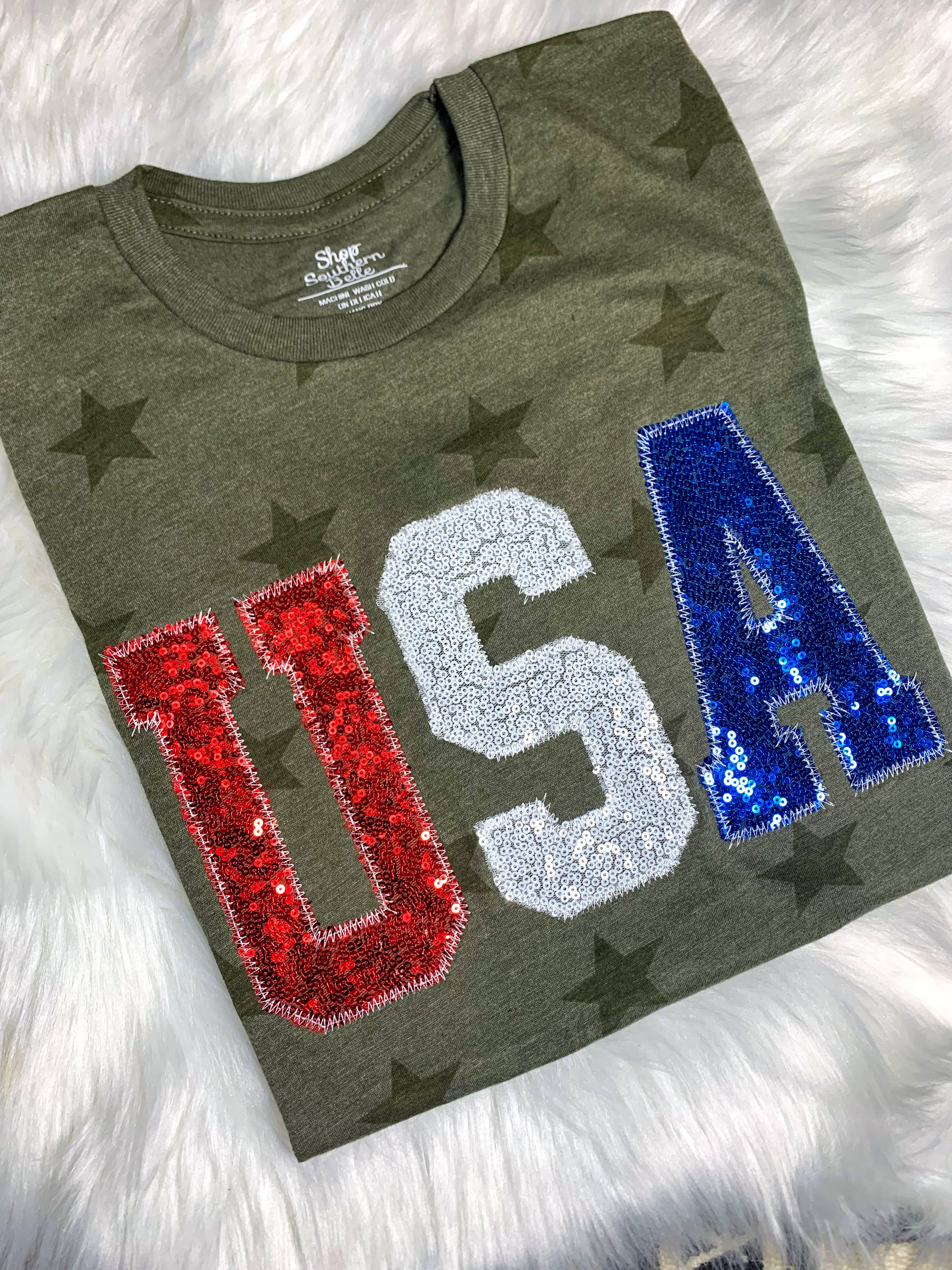 Green t-shirt with 'USA' in red, white, and blue sequins on a fluffy white surface