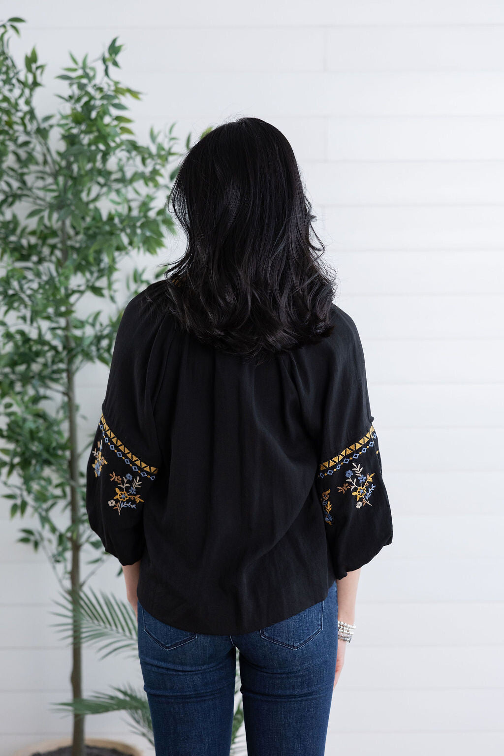 A woman viewed from behind wearing a black blouse with embroidered details on the sleeves and blue jeans, standing in front of a white paneled wall next to a green potted plant.