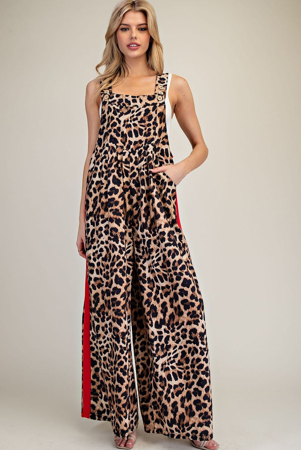 A woman modeling a leopard-print sleeveless jumpsuit with wide legs and red side panels, photographed against a plain light beige studio background.