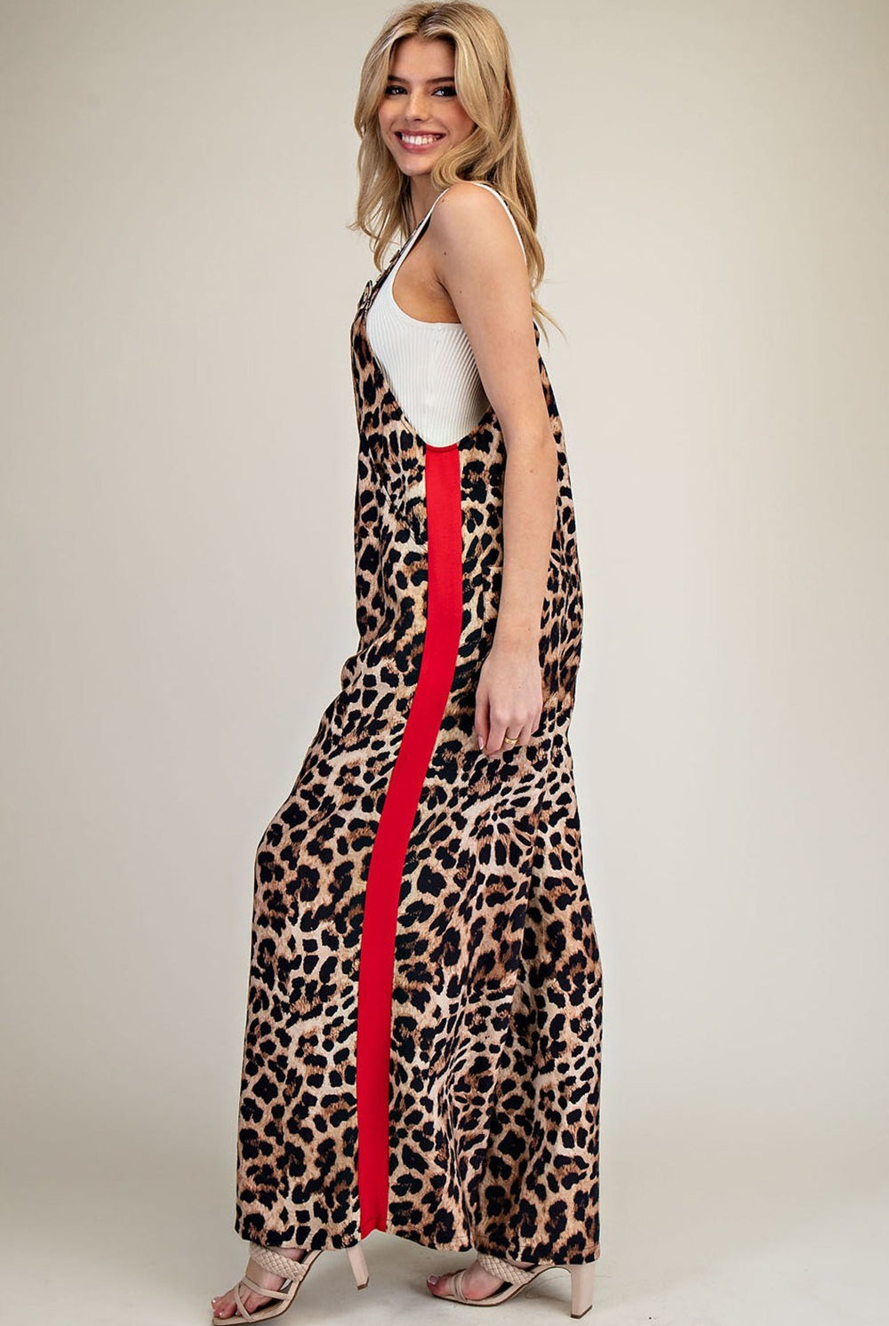 A woman modeling a sleeveless leopard-print maxi dress with a bold red side stripe, worn over a white tank top, photographed against a plain beige studio background.