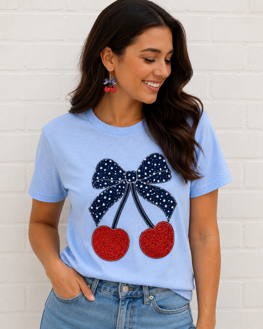 Woman wearing a light blue t-shirt with decorative bow and cherry design against a white brick wall.