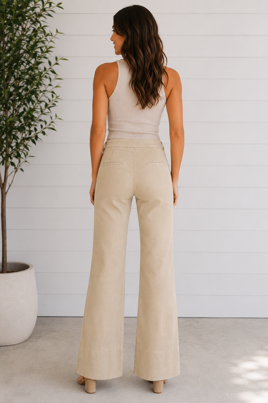 Woman wearing a beige sleeveless top and wide-leg pants standing in a minimalistic room with a plant.