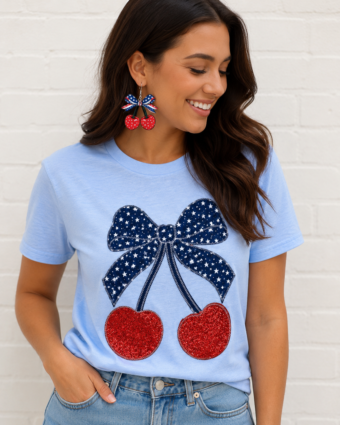 Woman wearing a light blue t-shirt with a decorative bow and cherries design, standing against a white brick wall.