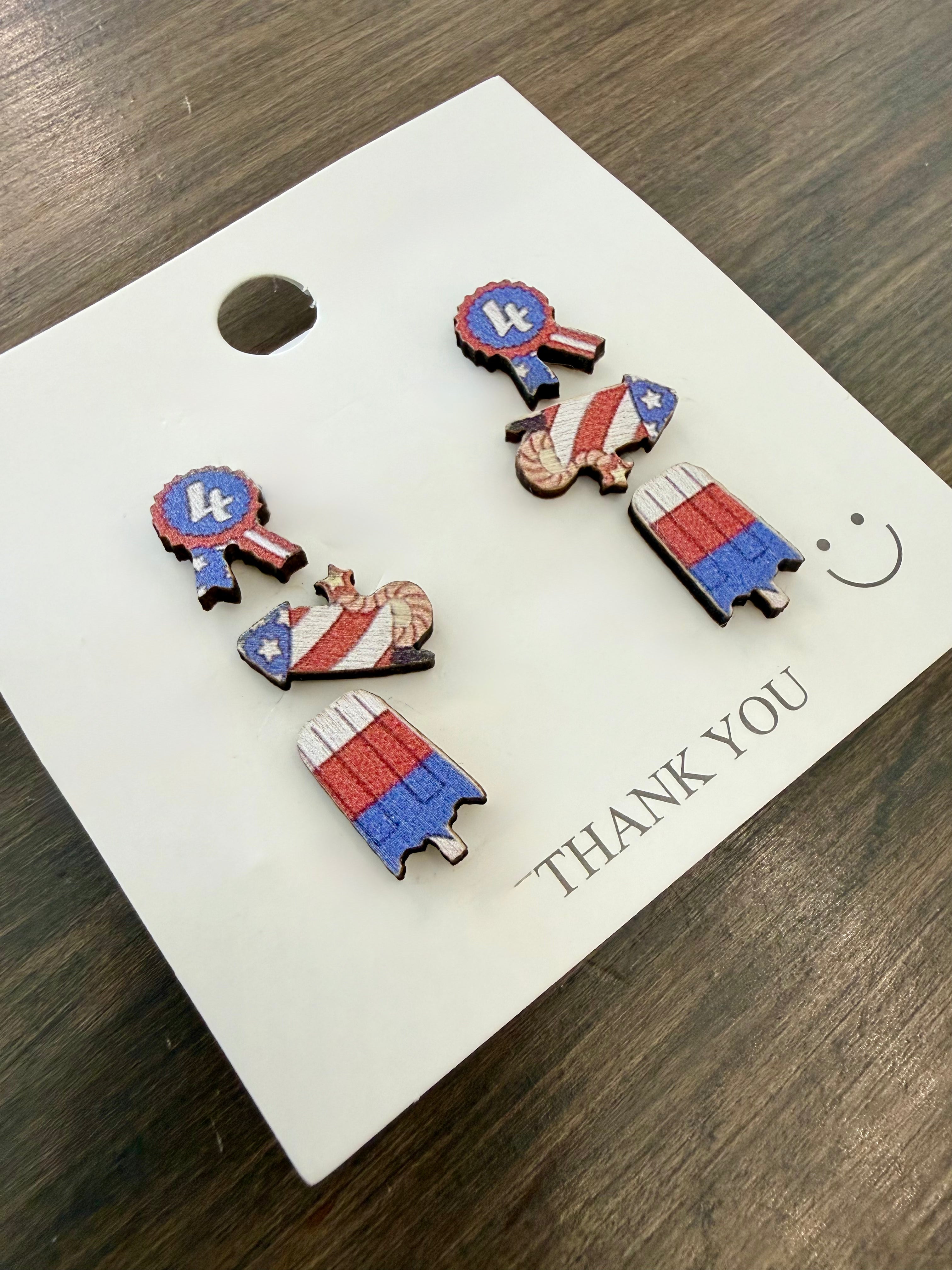 Set of 4th of July themed earrings on a 'THANK YOU' card with a smiley face on a wooden surface