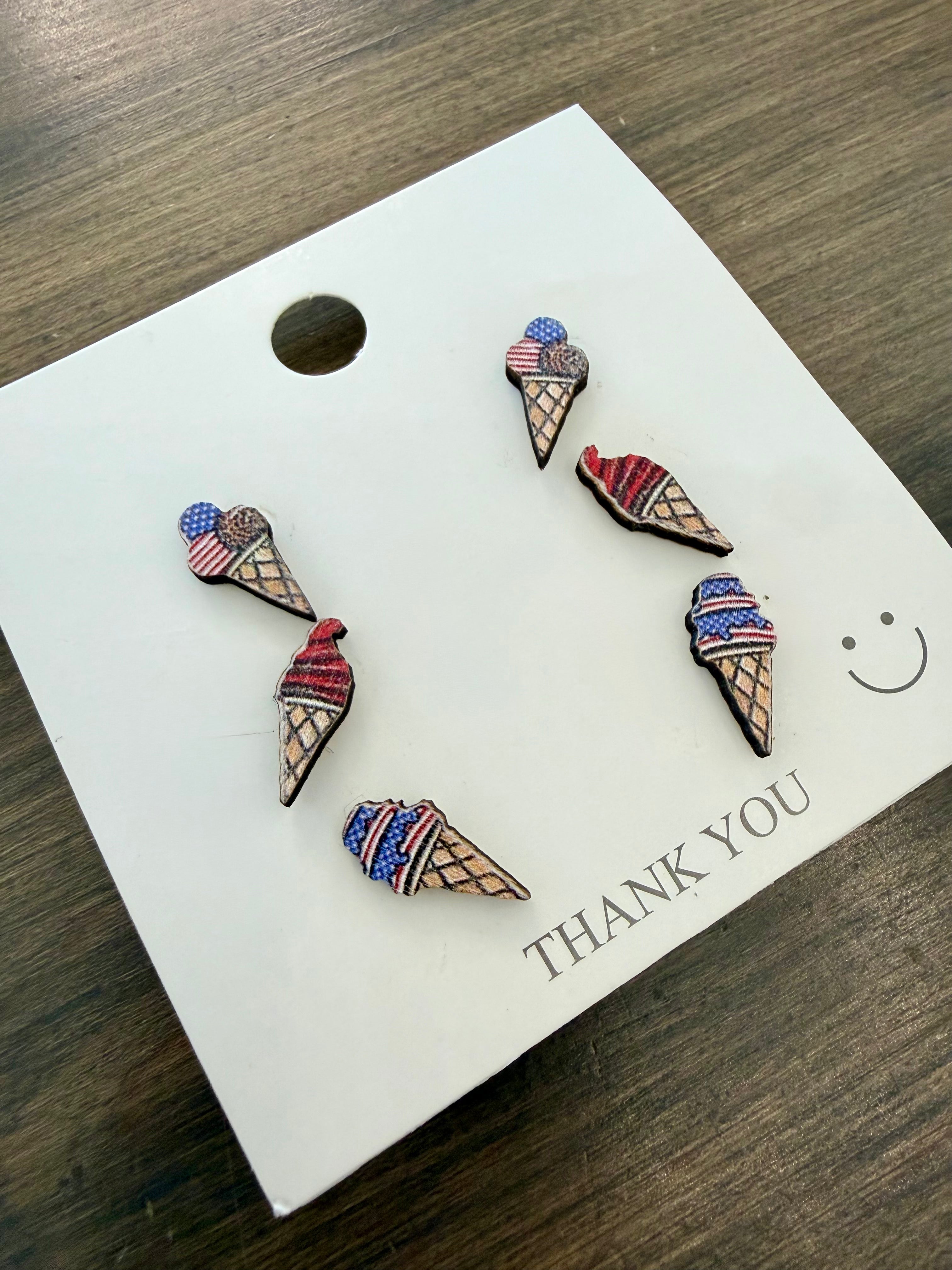 Ice cream cone-shaped earrings on a 'THANK YOU' card with a wooden surface background. Set of 4th of July themed earrings on a 'THANK YOU' card with a smiley face on a wooden surface
