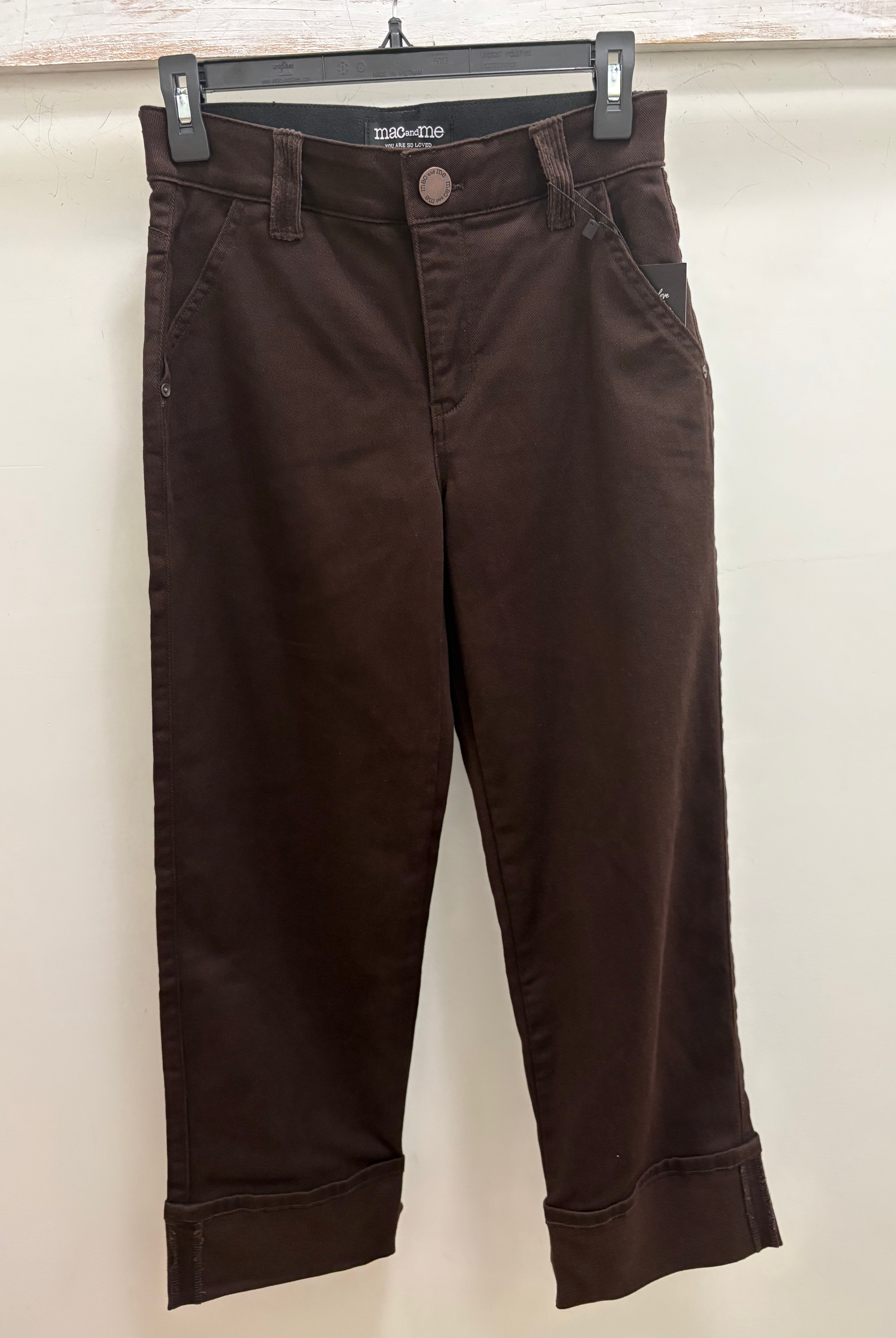 Dark brown cropped pants with rolled cuffs, hanging on a black hanger against a light beige wall.