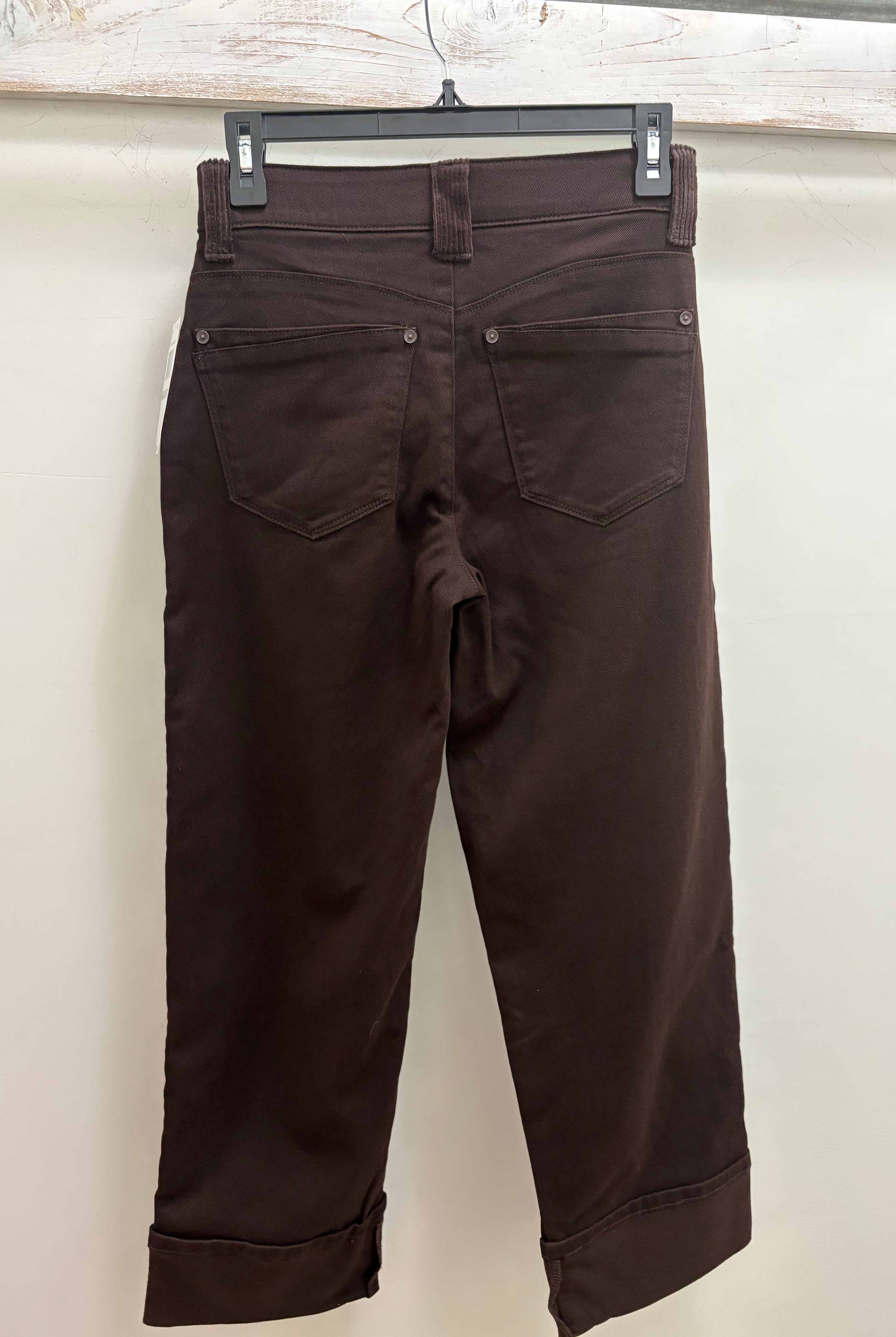 Dark brown cropped pants shown from the back, hanging on a black hanger against a plain off-white wall.