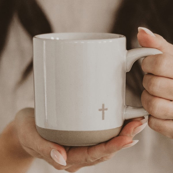 White mug with a cross symbol held by two hands against a neutral background