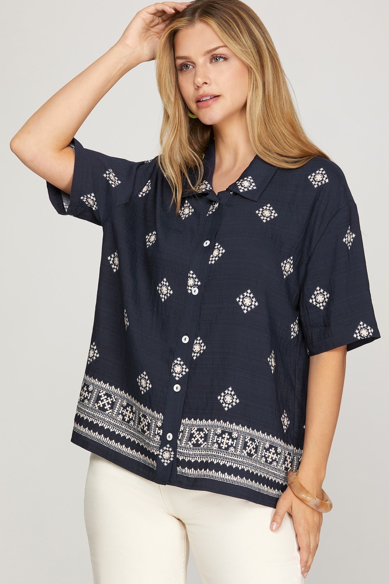 A woman wearing a navy blue short-sleeve button-up shirt with white geometric and tribal-style prints, paired with white pants, against a light gray studio background.
