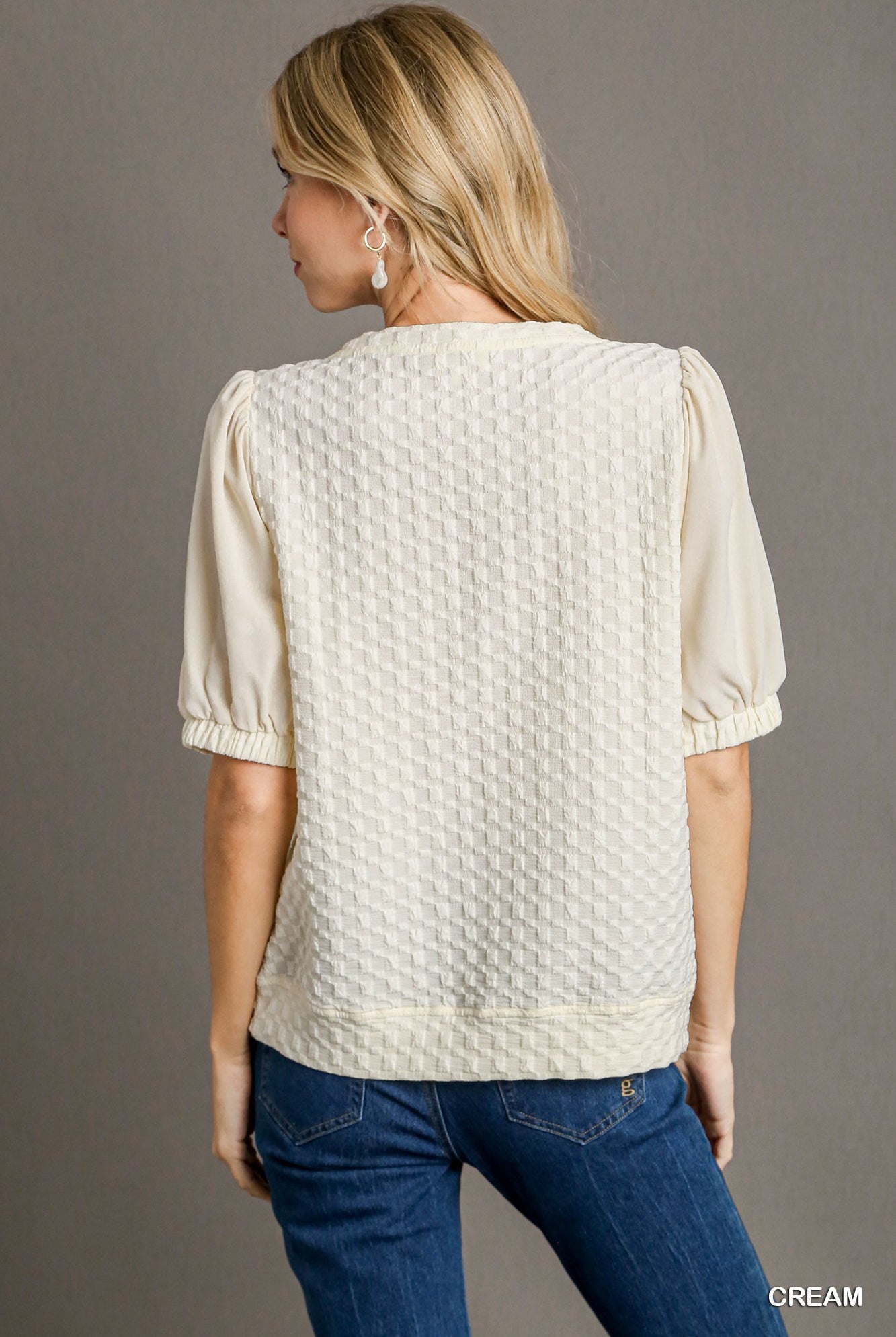Woman wearing a cream textured blouse and blue jeans on a gray background