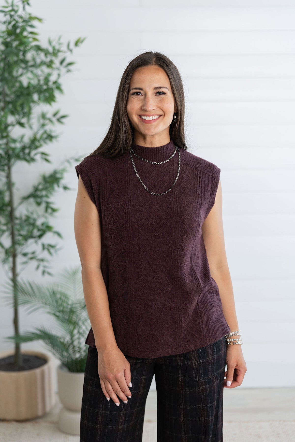 A woman wearing a sleeveless, mock-neck knit top in a deep plum purple color, paired with dark plaid pants, standing in front of a white paneled wall with potted plants.