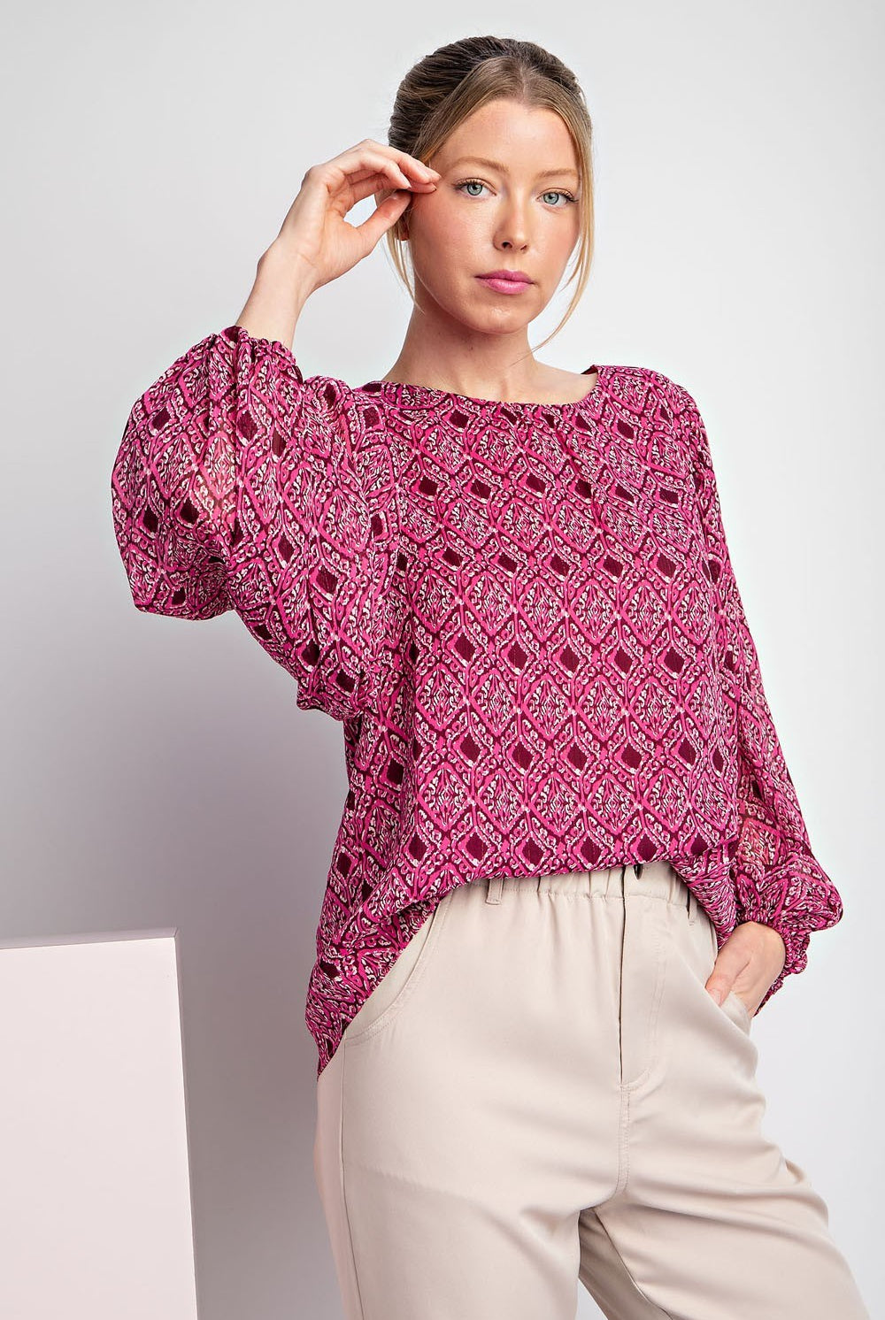 Woman wearing a pink patterned blouse and beige pants against a white background