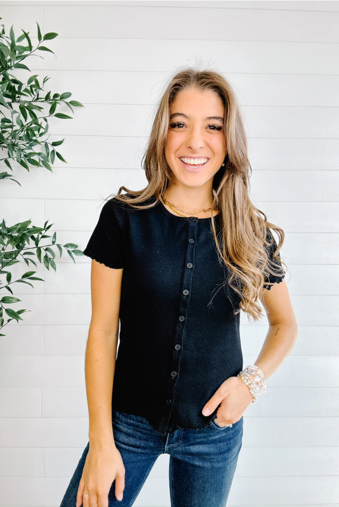 Woman wearing a black button-up shirt and jeans standing against a white brick wall with a plant to the left.