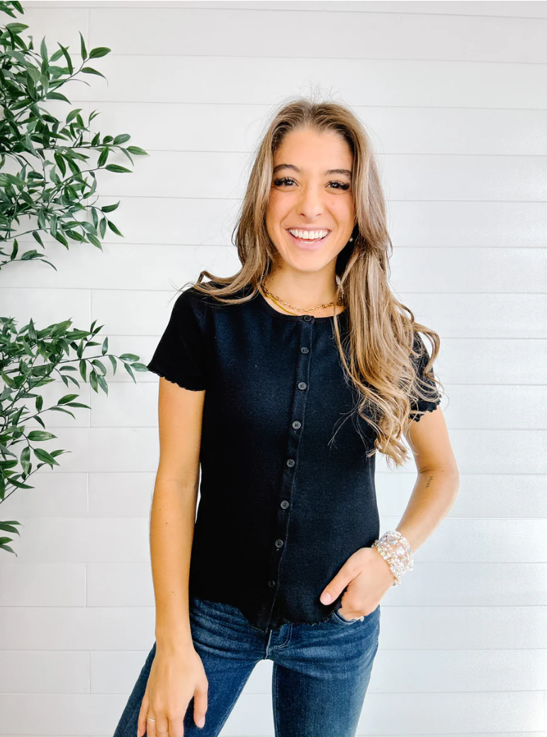 Woman wearing a black button-up shirt and jeans standing against a white brick wall with a plant to the left.