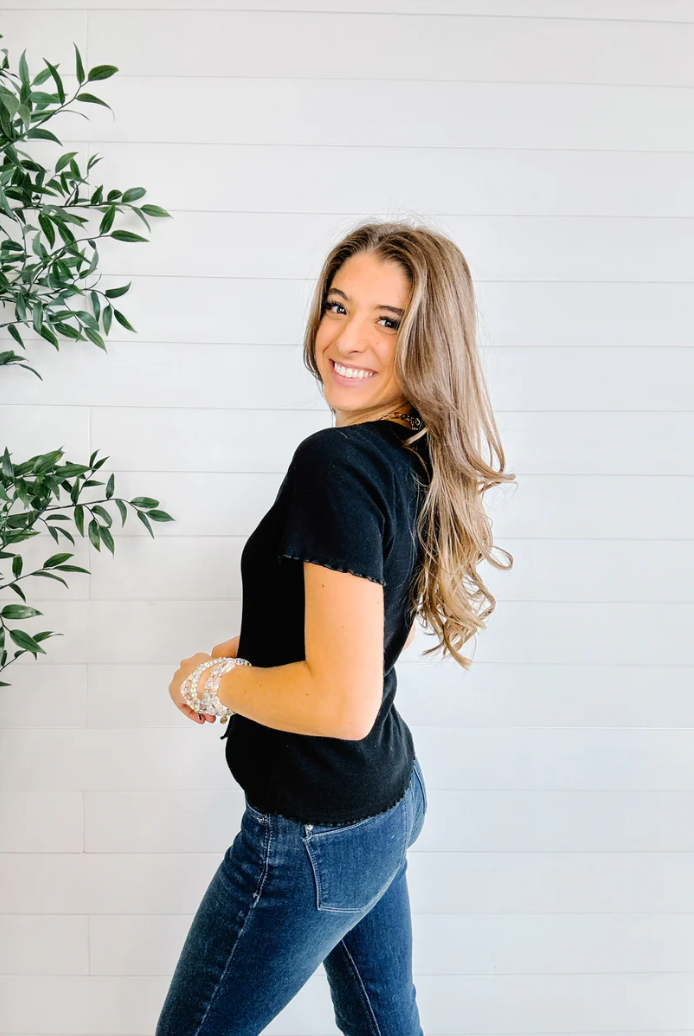Woman wearing a black top and blue jeans standing against a white wall with a plant to the left.