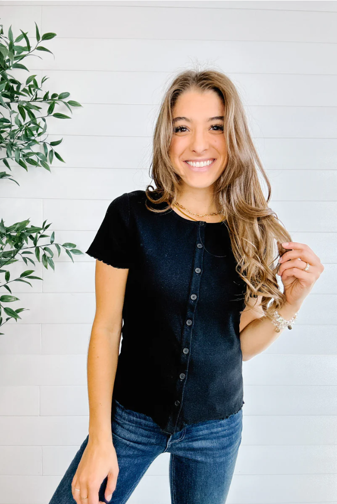 Woman wearing a black button-up shirt and blue jeans standing against a white brick wall with a plant to the left.