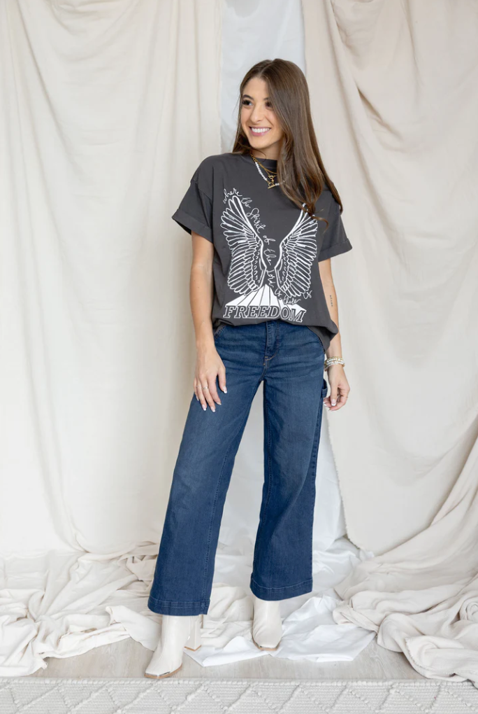Woman wearing a graphic t-shirt and blue jeans standing against a white curtain.