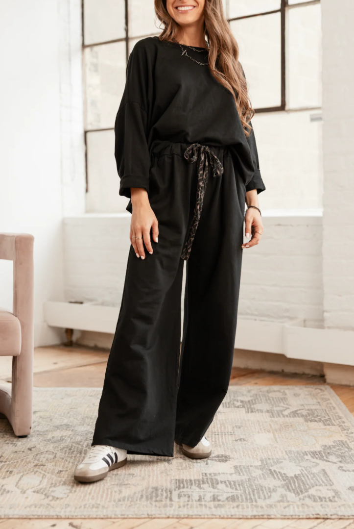 Woman wearing a black outfit with wide-leg pants in a room with large windows.