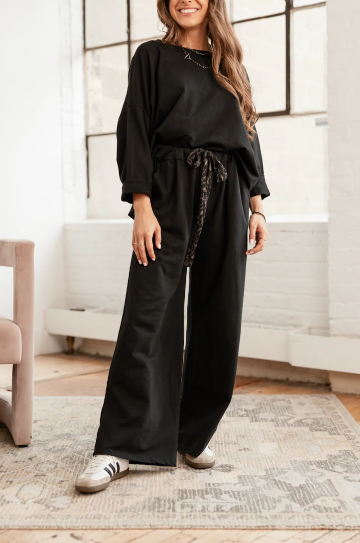 Woman wearing a black outfit with wide-leg pants in a room with large windows.