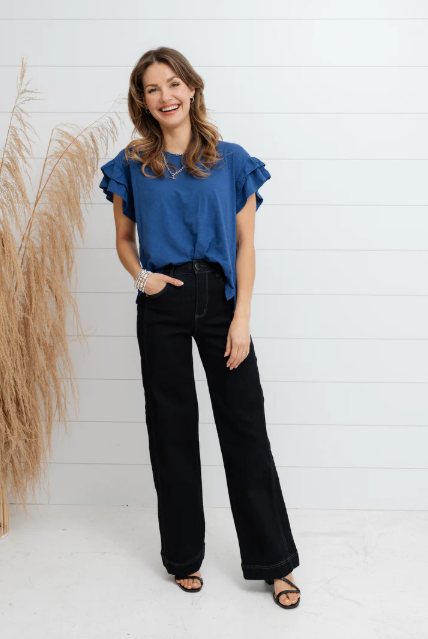 Woman wearing a blue top and black pants standing against a white wall with decorative elements.
