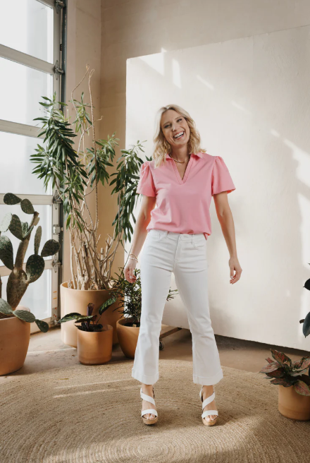 Woman in a pink shirt and white pants standing in a room with plants.