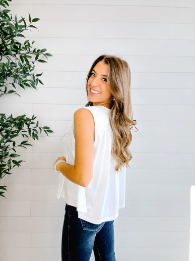 Woman wearing a white sleeveless top and blue jeans standing against a white wall with a plant to the left.