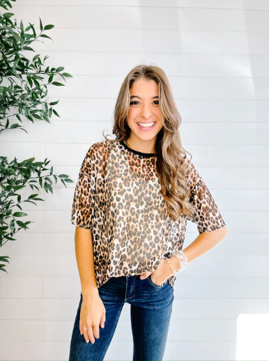 Woman wearing a leopard print top and blue jeans standing in front of a white wall with a plant.