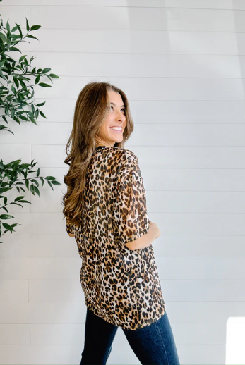 Woman wearing a leopard print shirt standing next to a plant against a white wall