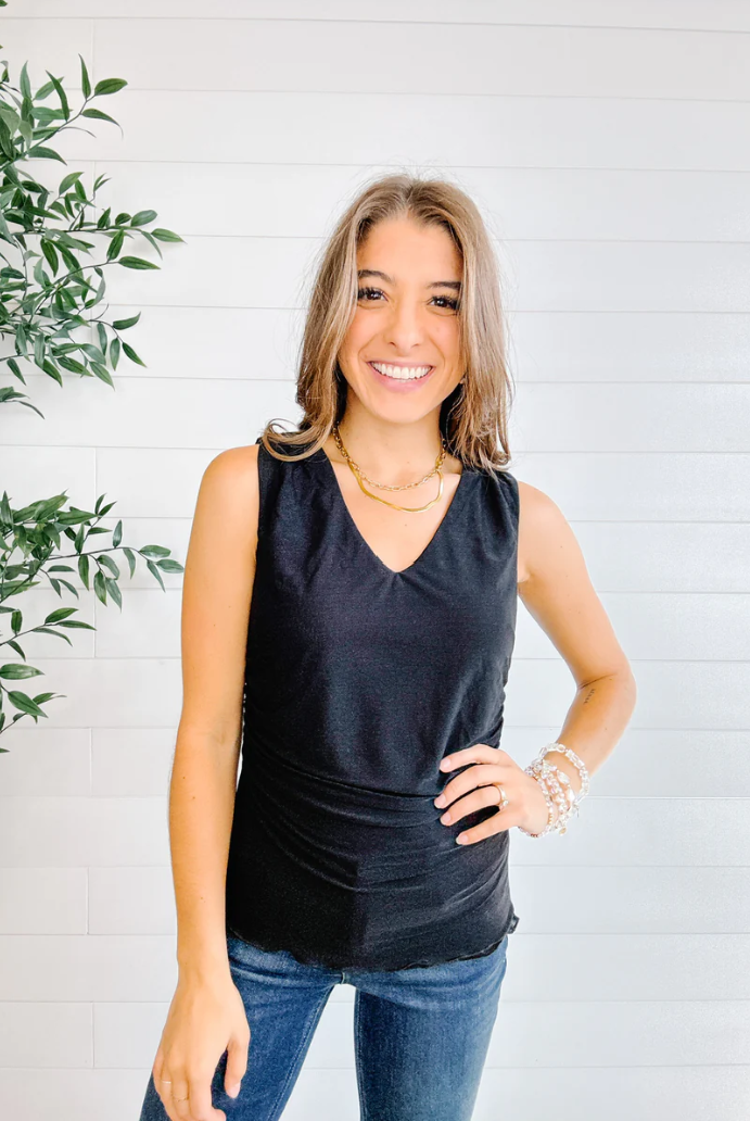Woman wearing a black sleeveless top and blue jeans standing against a white wall with a plant to the left.
