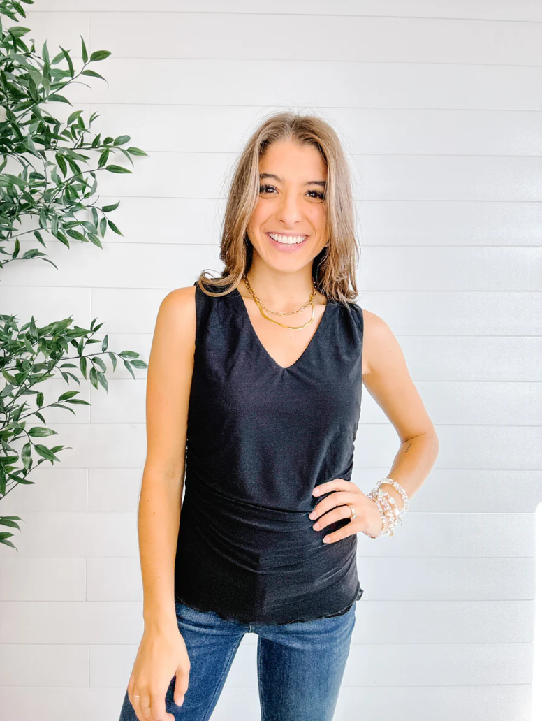 Woman wearing a black sleeveless top and blue jeans standing against a white wall with a plant to the left.
