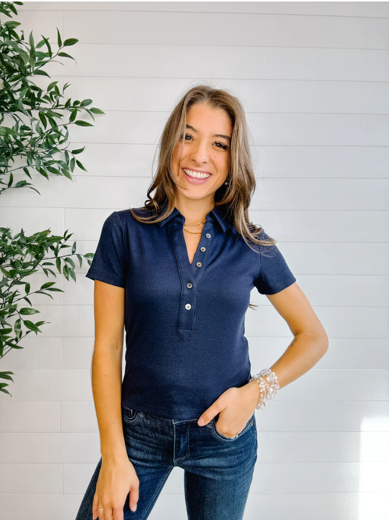 Woman wearing a navy blue polo shirt and jeans standing against a light wooden panel background with a plant to the left.