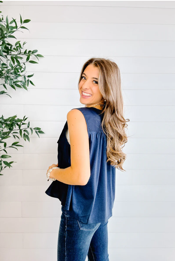 Woman wearing a denim sleeveless top and jeans standing against a white wall with a plant to the left.
