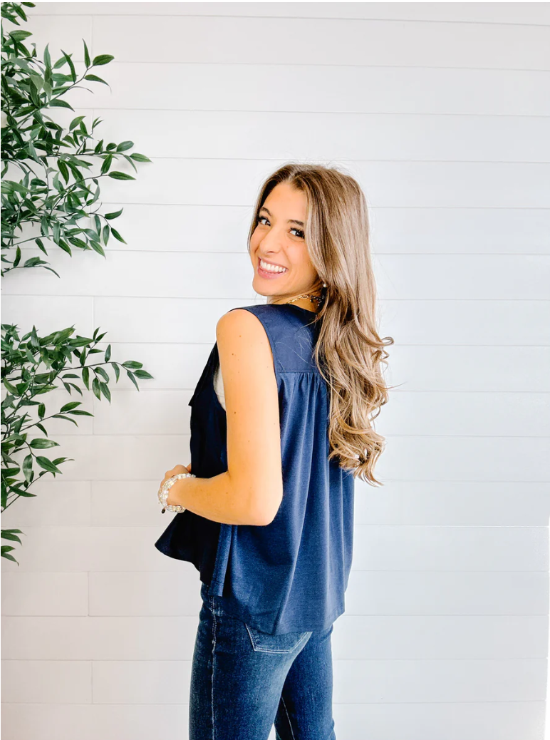 Woman wearing a denim sleeveless top and jeans standing against a white wall with a plant to the left.