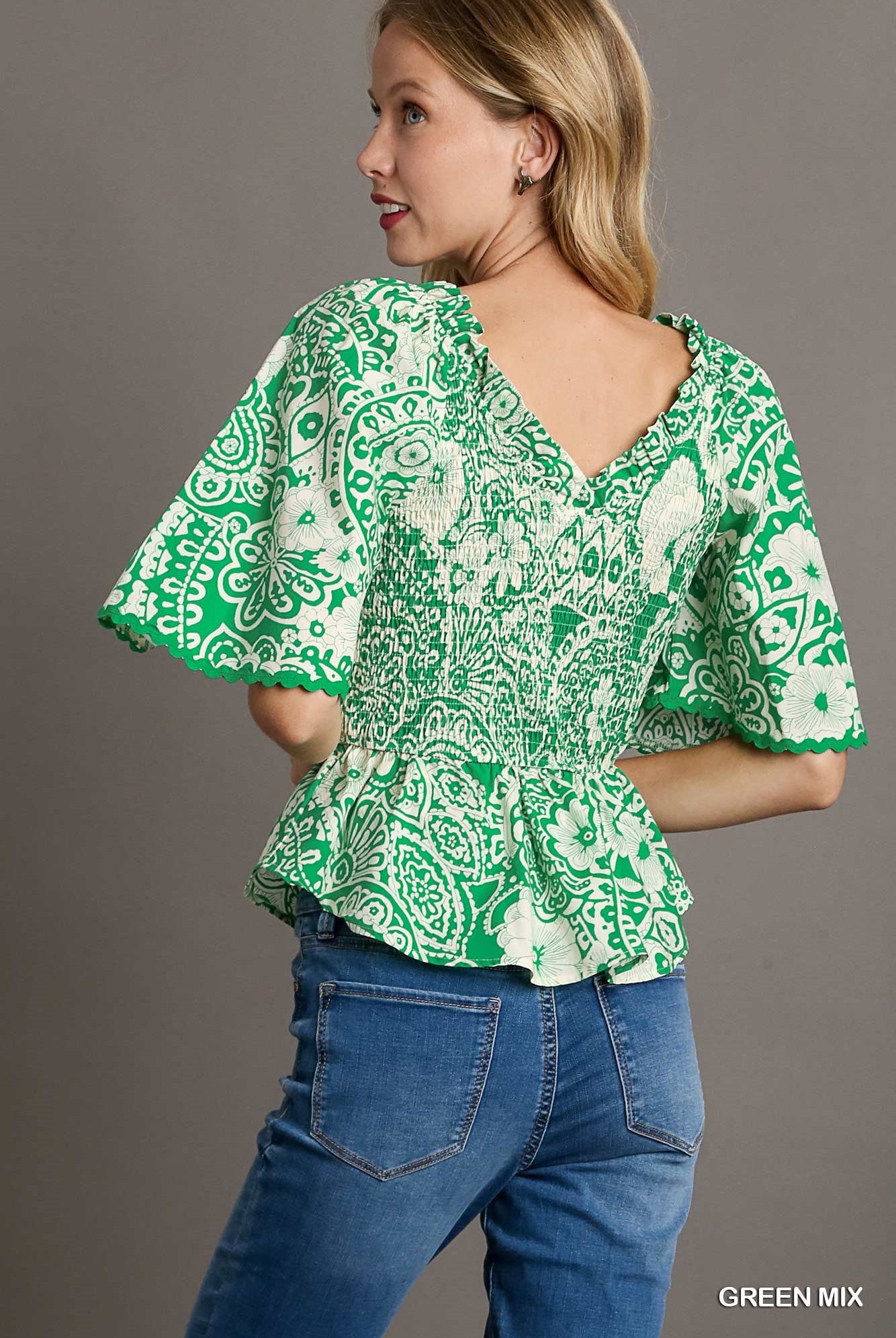 Woman wearing a green and white patterned blouse with blue jeans against a gray background