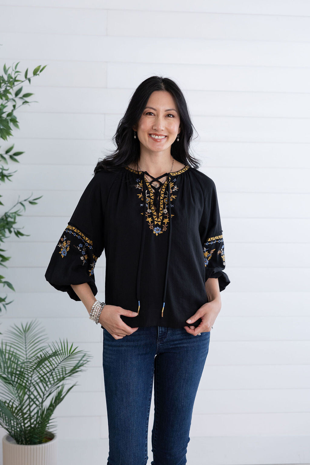 A woman wearing a black long-sleeve blouse with gold and blue embroidered details on the neckline and cuffs, paired with dark blue jeans, standing against a white shiplap wall with potted plants.