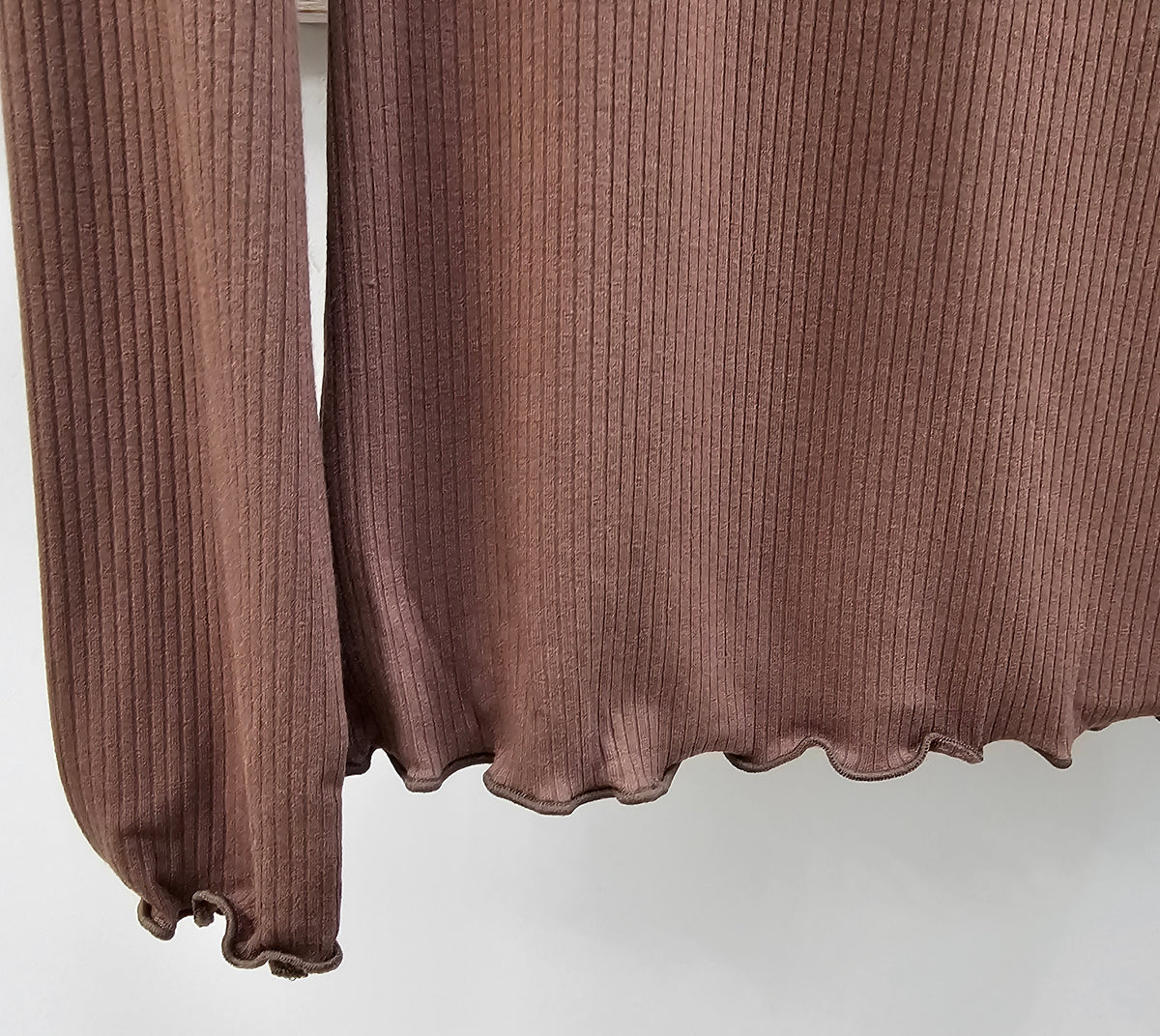 Close-up of a ribbed long-sleeve top in a muted dusty rose or mauve-brown color, showing the sleeve cuff and ruffled hem against a plain light background.