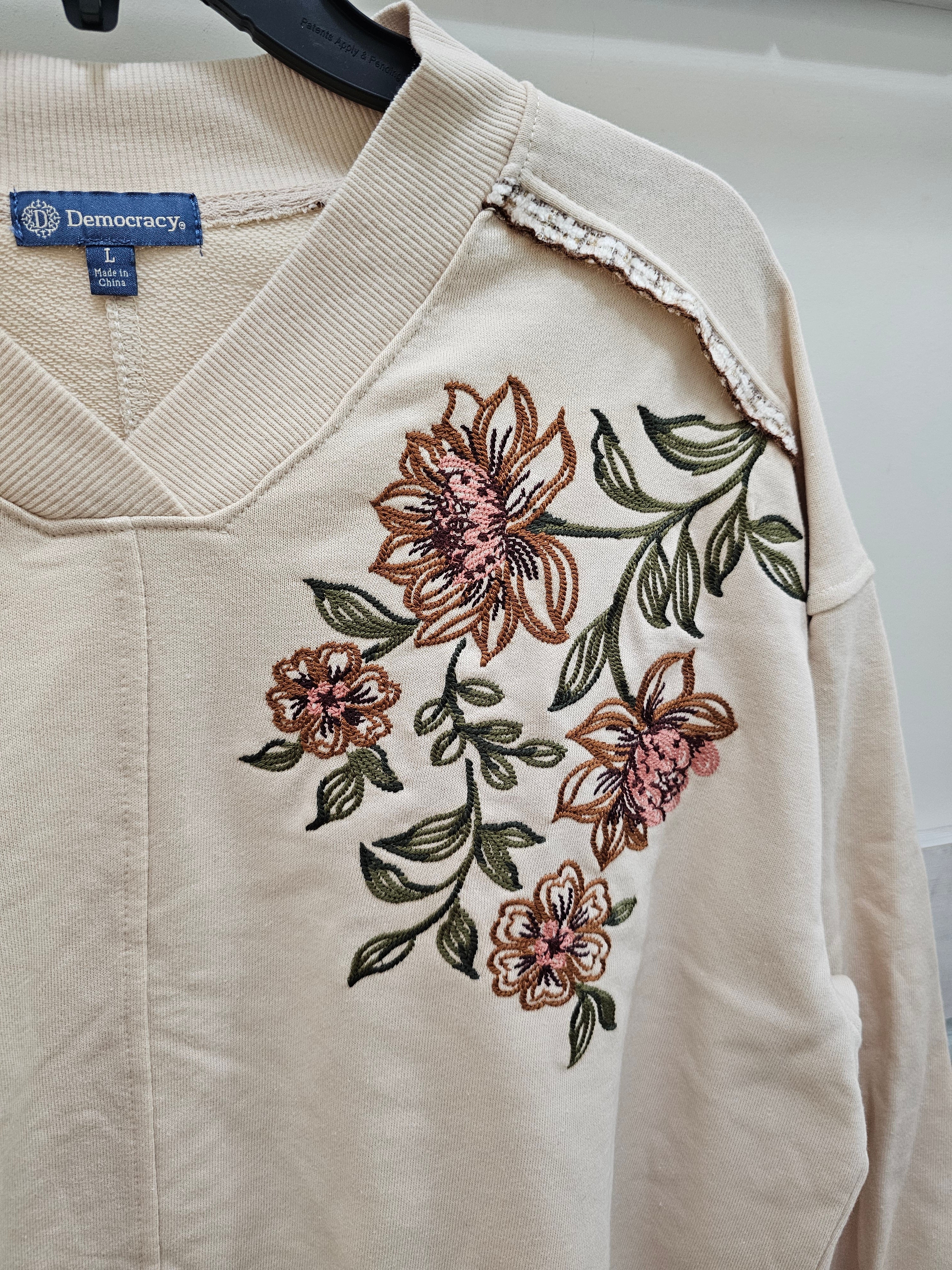A close-up of a cream-colored long-sleeve V-neck sweater hanging on a black hanger. The sweater features detailed floral embroidery in brown, pink, and green on the front, with a lace-trimmed shoulder cutout.