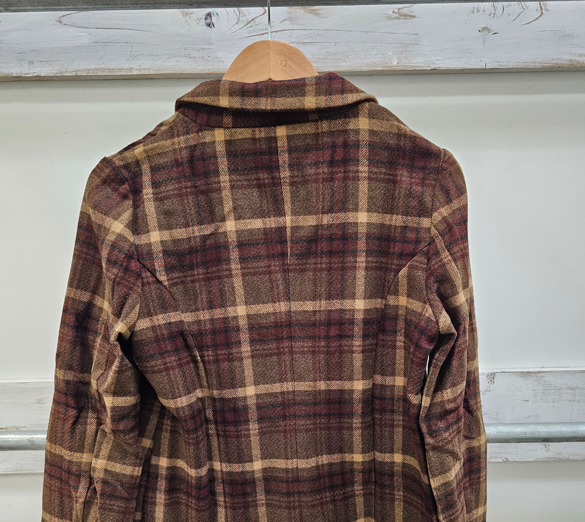 A vintage plaid flannel shirt jacket hanging on a wooden hanger against a white wall with rustic wooden beams.