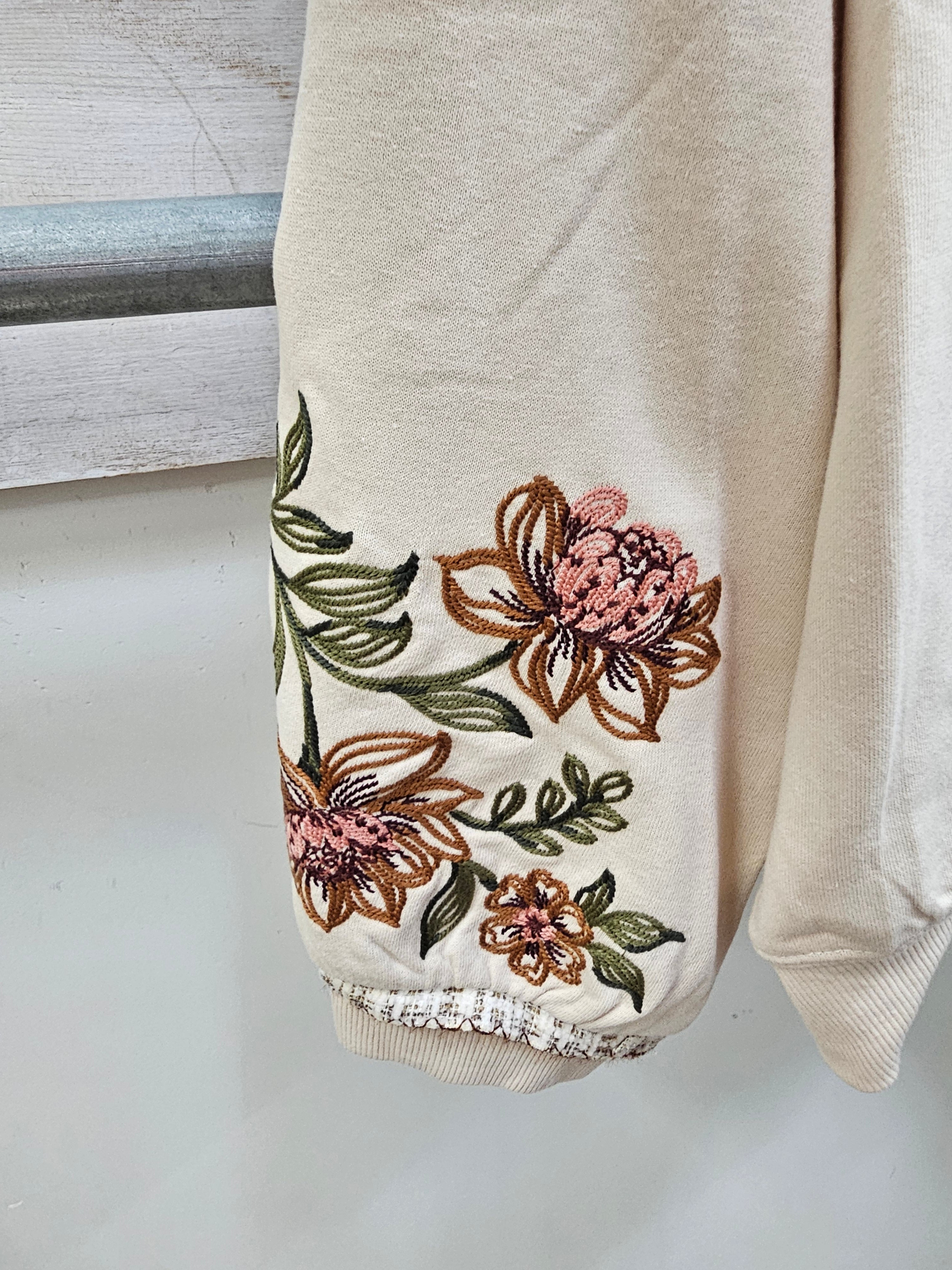 Close-up of a cream-colored sweatshirt sleeve featuring embroidered pink and brown flowers with green leaves, finished with a frayed ribbed cuff.