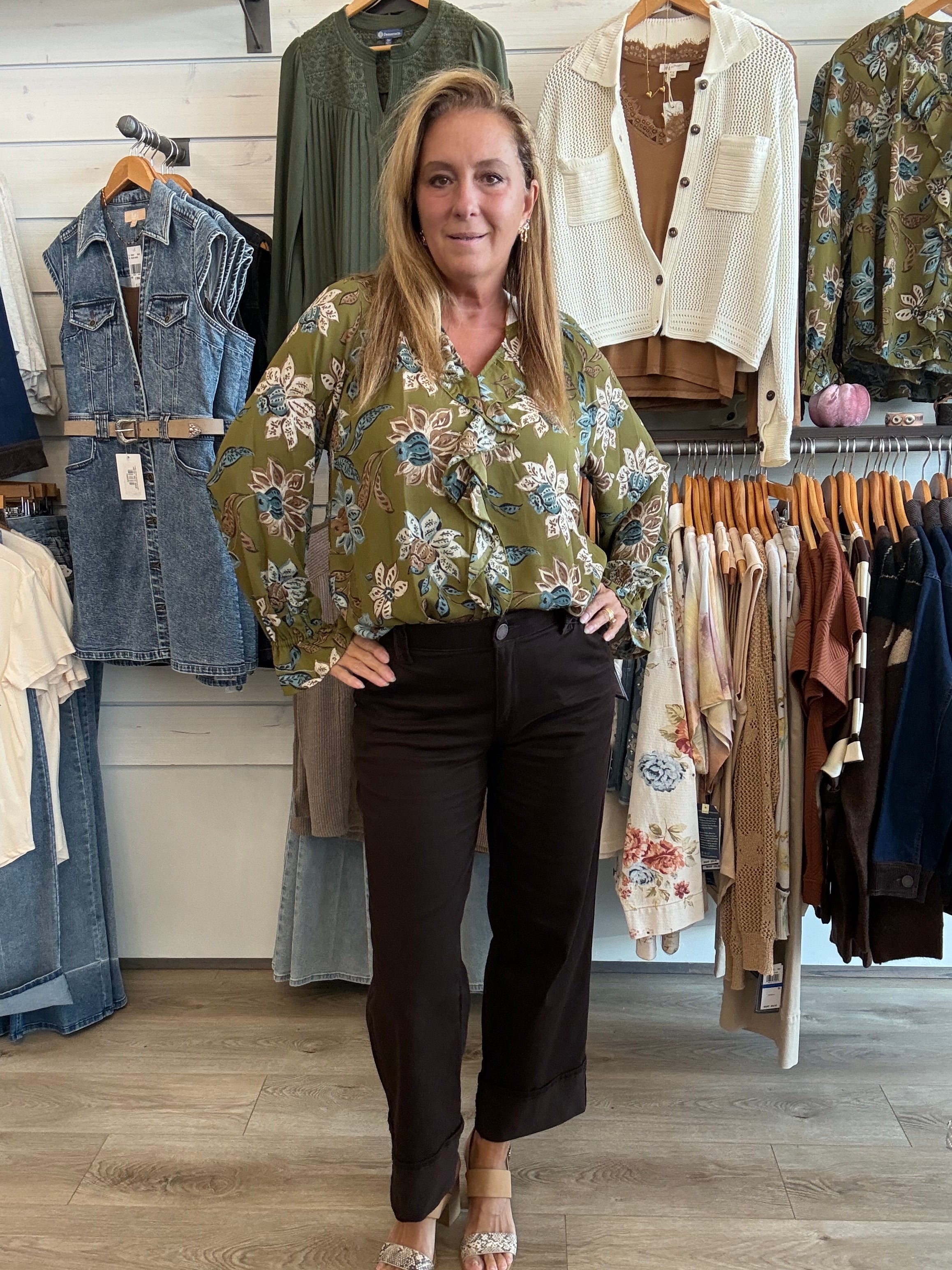 A woman stands in a clothing boutique wearing a sage green floral long-sleeve blouse and dark brown cropped pants. Clothing racks and shelves are visible behind her.