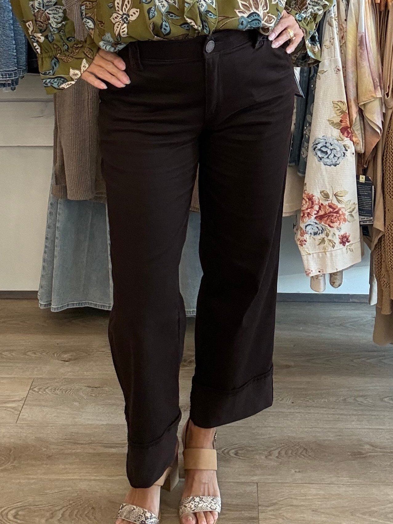 A person wearing dark brown cropped pants with rolled cuffs, paired with a floral blouse and beige heeled sandals, standing in a clothing store on a wood floor.