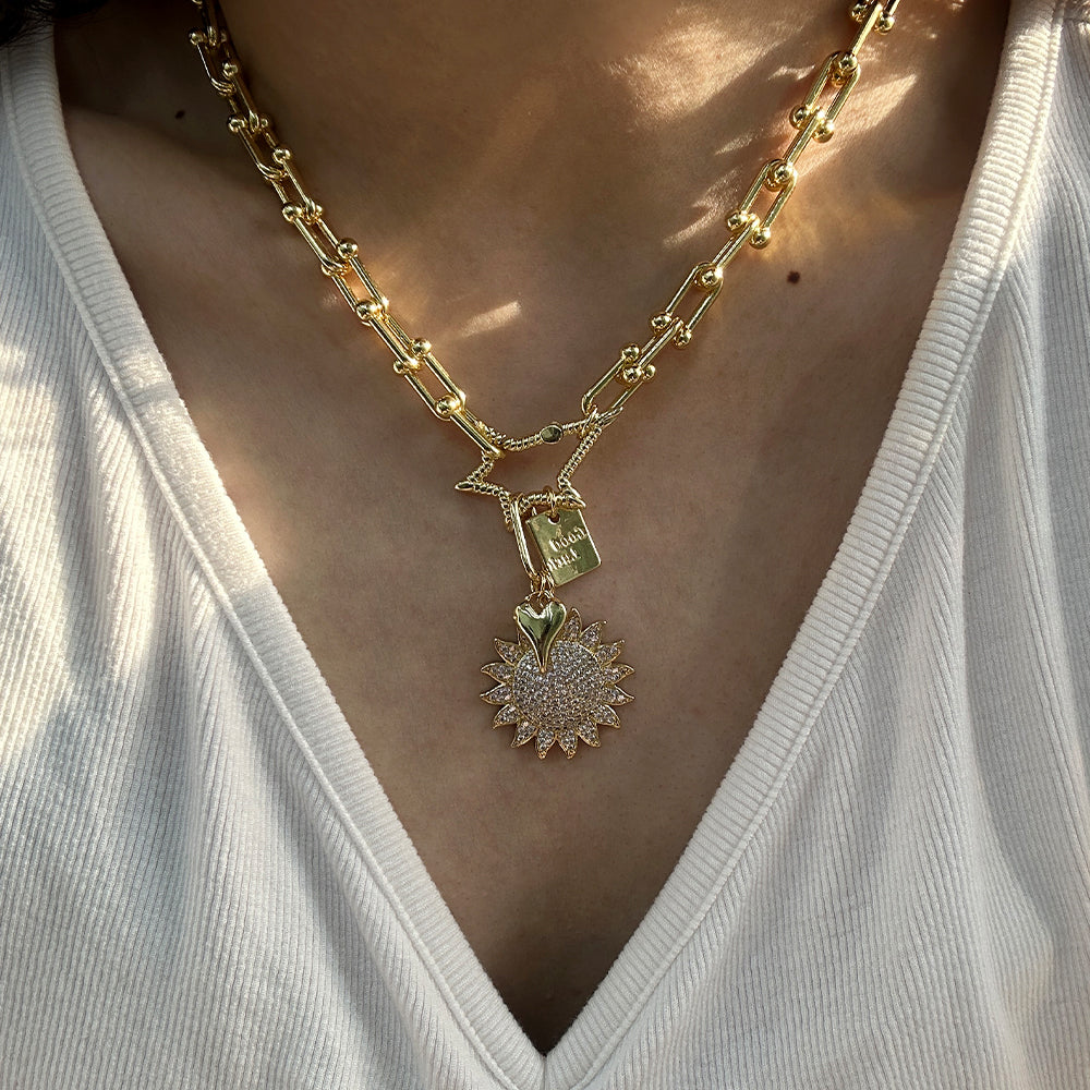 A close-up of a person wearing a gold chain necklace with a star-shaped connector and a sunburst pendant adorned with small clear stones, styled with a white ribbed V-neck top.