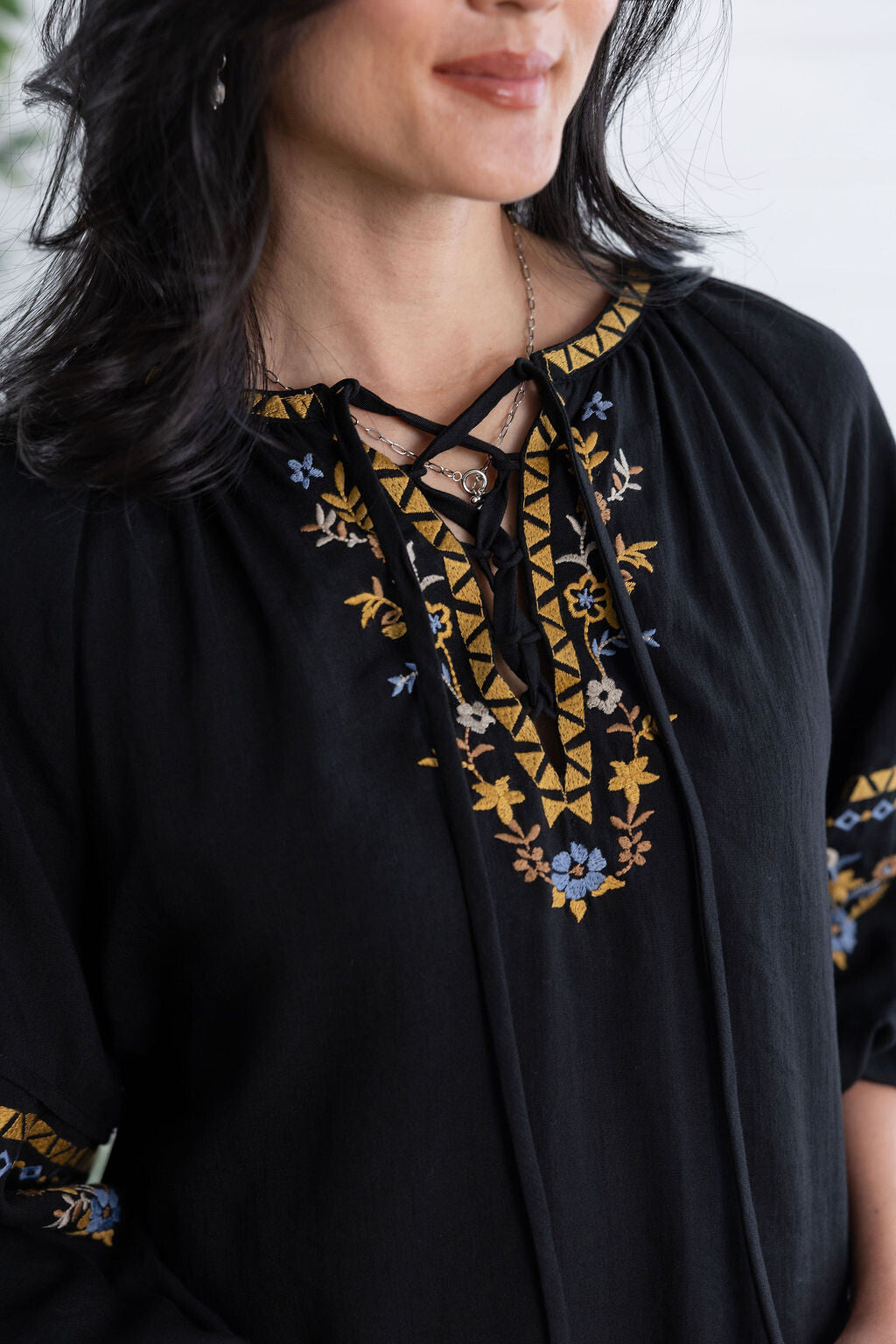 A close-up of a woman wearing a black embroidered tunic top with lace-up neckline details in gold, blue, and white, set against a bright, softly blurred background.