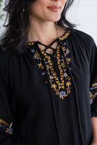 A close-up of a woman wearing a black embroidered tunic top with lace-up neckline details in gold, blue, and white, set against a bright, softly blurred background.