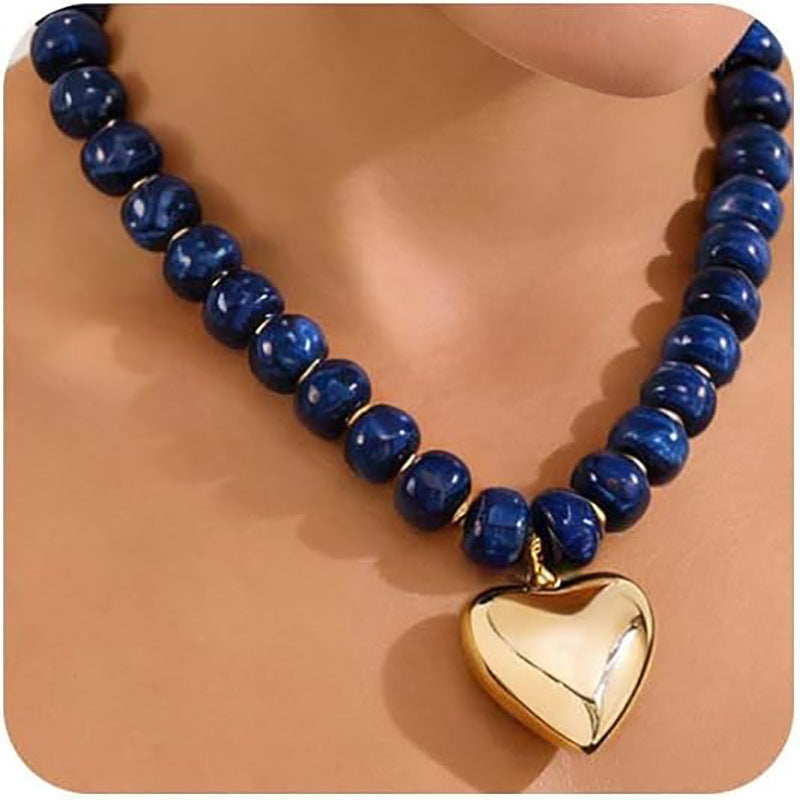 A beaded necklace with polished dark blue round beads and a shiny gold heart-shaped pendant, displayed on a mannequin neck against a warm beige background.